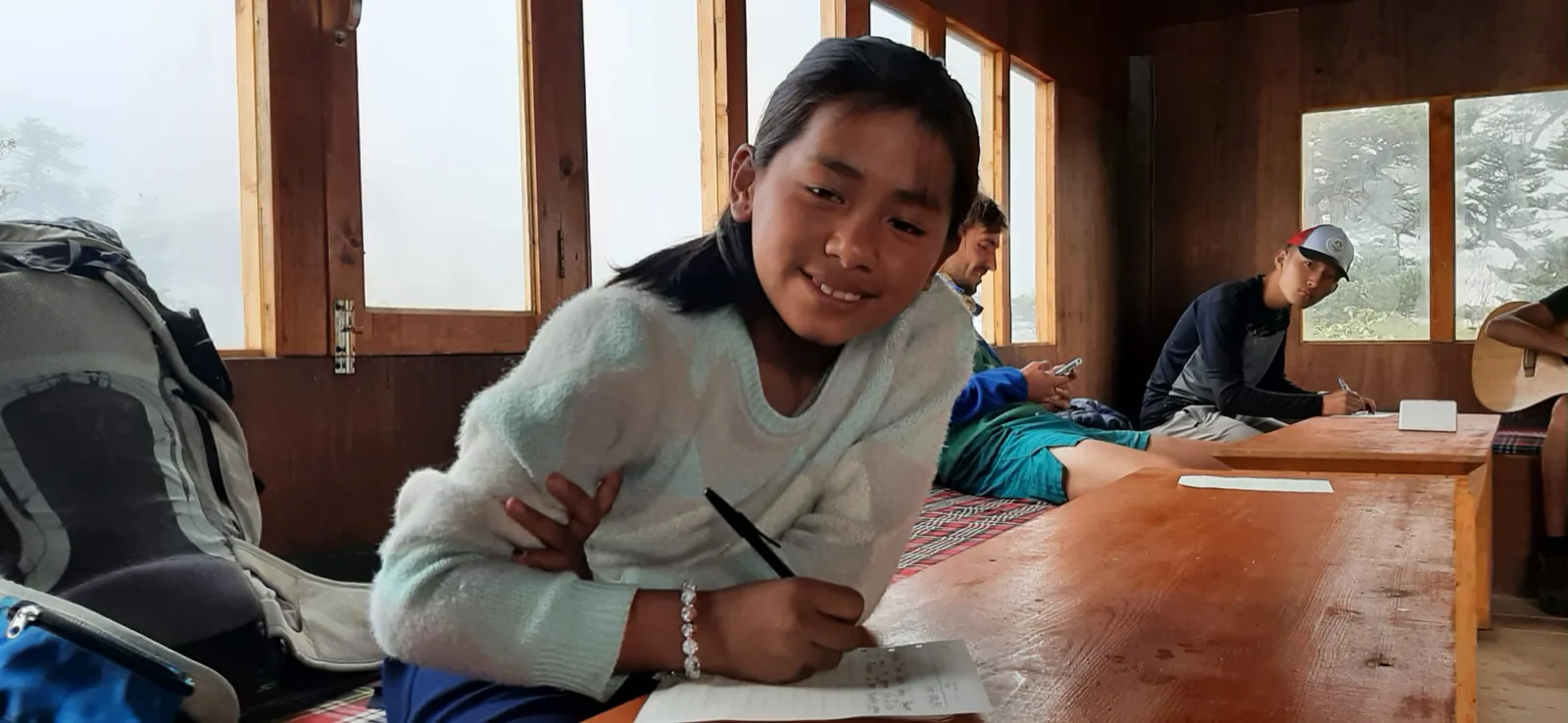 Une élève souriante écrit dans son cahier, assise à une longue table en bois