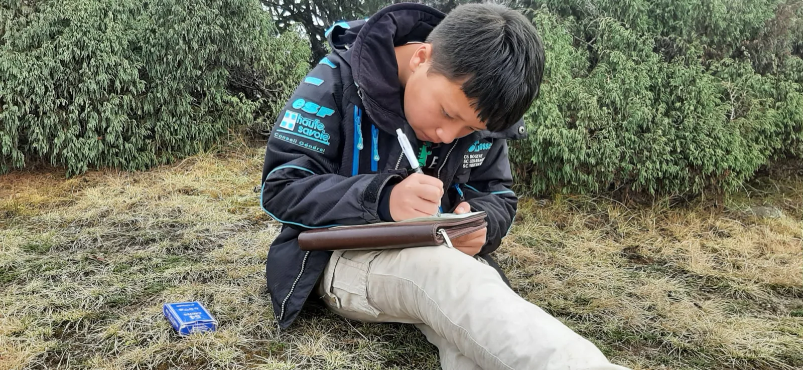 Un garçon assis dans l'herbe écrit dans un carnet posé sur ses genoux